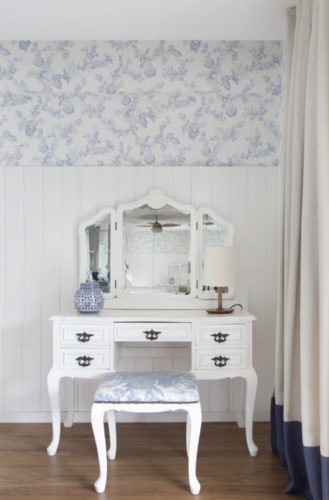 Decoração clássica; meia parede em lambri branco, e pra cima, papel de parede floral claro, na frete; penteadeira branca 
