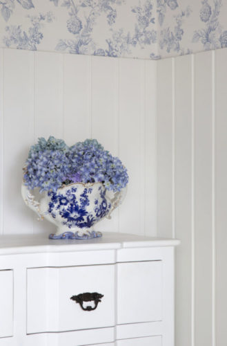 Decoração clássica. Lambri branco em meia parede, pra cima , papel de parede com fundo branco e flores azuis. Em cima de uma cômoda branca, um vaso de porcelana azul e branco com hortênsias azuis 