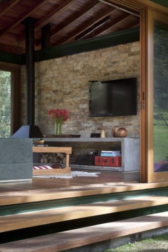 Sala de uma casa no campo, parede em pedra, teto em madeira e rack em cimento.