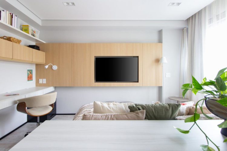 Apartamento de 30m2 de uma jovem solteira, em frente cama, que faz as vezes de sofá, um painel de madeira embute a tv e alguns armários, ao lado, uma bancada de estudos