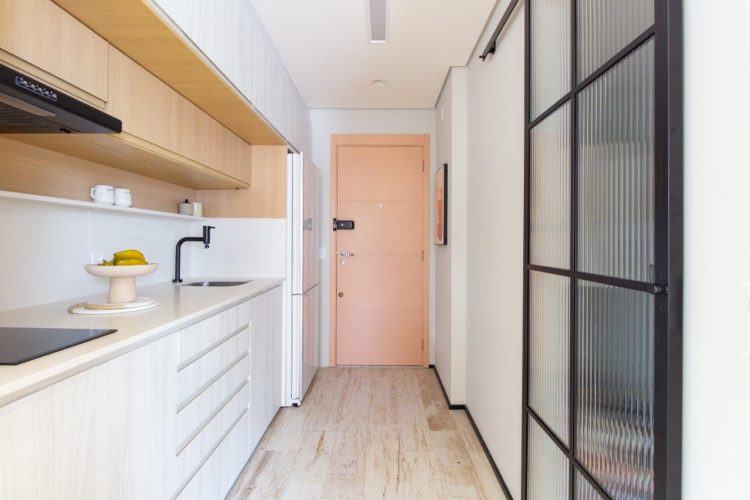 Apartamento de 30m2 de uma jovem solteira, entrada com a porta rosa, e de um lado a bancada de cozinha, com armários na parte de baixo .Cuca e cooktop 