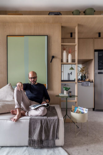 Arquiteto Leandro Neves sentado no sofá cinza com parede de madeira ao fundo.
