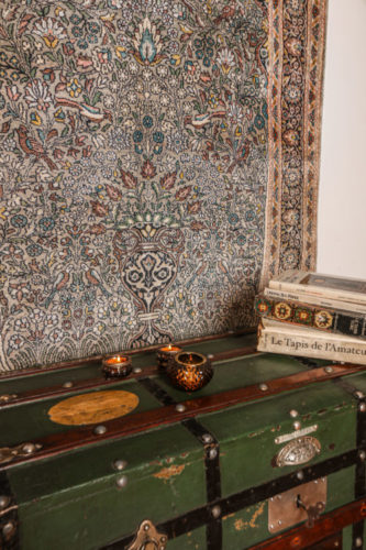 Tapete Kayseri - turco pendurado na parede e em frente, um baú verde com livros sobre tapetes em cima.
