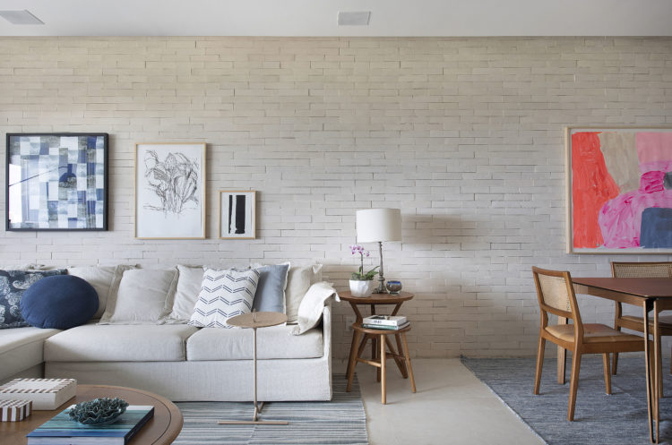 Living decorado em tons claros. Sofá na cor off-white e parede ao fundo revestida com tijolinhos pintados de branco.