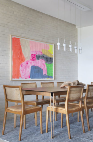 Ambiente de sala de jantar com mesa e cadeiras em madeira, parede revestida de tijolinhos e um quadro colorido