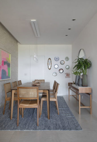 Ambiente de mesa de jantar. Mesa e cadeiras em madeira, ao fundo, uma composição de pratos na parede.