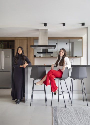 As arquitetas Luana Bergamo e Luiza Mesquita, uma em pé ao lado do balcão que integra a cozinha e a outra sentada em uma das banquetas