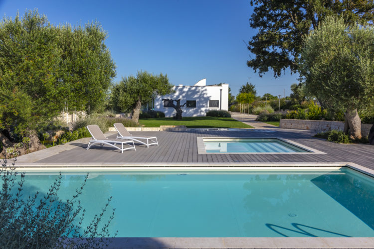 Deck com duas piscinas, uma pequena e outra grande,, jardim ao lado com oliveiras, e ao fundo uma casa branca