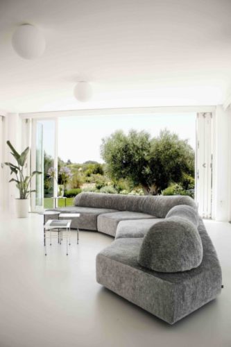 Decoração contemporânea, com uma sofá cinza grande, formato orgânico, janelões abertos e a natureza integrada. 