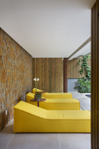 Área da piscina com três chaises na cor amarela e na parede de fundo, revestimento em 3d com placas de imitando aço corten.