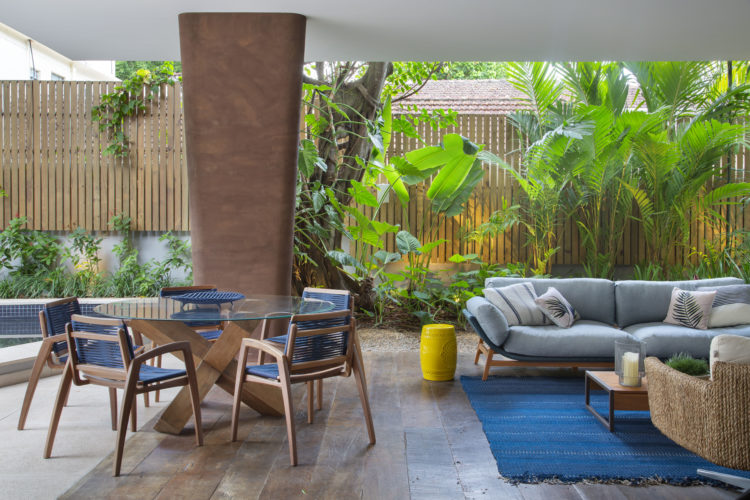 Sala integrada com o espaço externos. Jardim ao fundo, mesa redonda com cadeiras, tapete azul e um sofá azul claro decoram o ambiente.