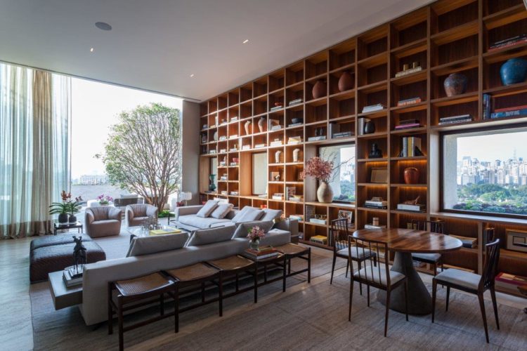 Grande living, claro , com mobiliário de designers brasileiros e na extensão de parede de fundo, uma grande estante com recortes para algumas janelas, que mais se parecem com quadros.