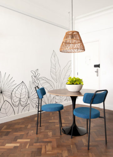 Desenhos na cor preta e branca de folhagens tropicais, na parede fundo. Mesa redonda com duas cadeiras azuis e um pendente de palha em cima