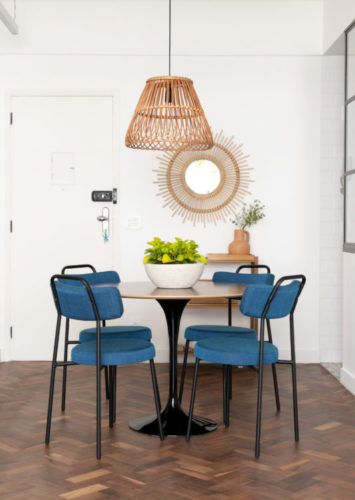 Mesa de jantar redonda, com cadeiras azuis, pendente em palha, e ao fundo espelho redonda também em palha 