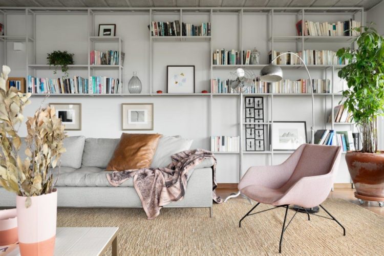 Estante de ferro ao funda na parede, com livros e objetos, sofá cinza em frente, poltona rosa ao lado 