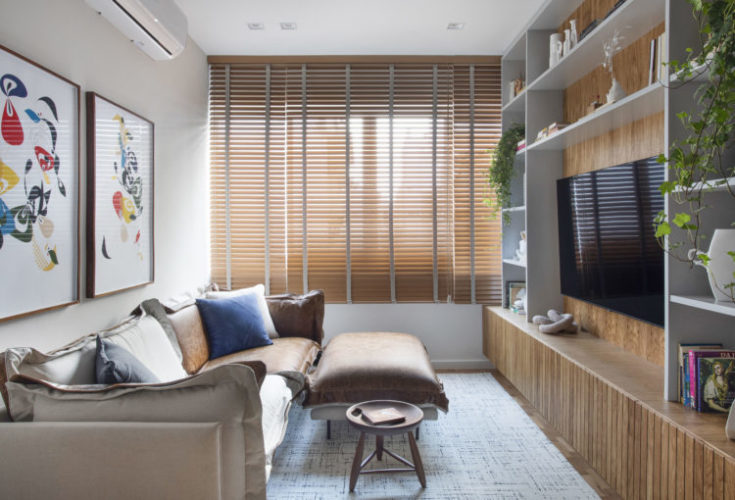Sala em formato de retângulo, com persianas horizontais em madeira, sofá em couro e linho, em frente a estante em laca branca e fundo em madeira