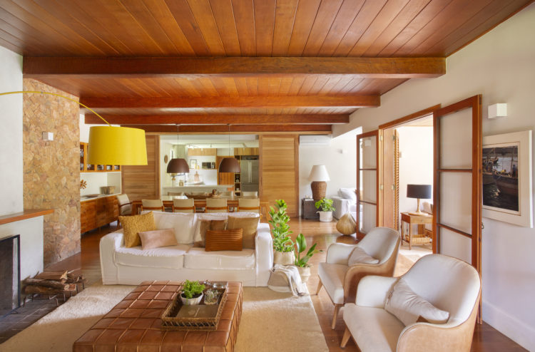 Sala em uma casa de campo, decorada em tons claros, com teto em madeira e parede de pedra ao lado da lareira.