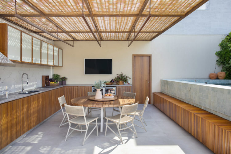 Área externa de uma cobertura duplex, piscina em cima de um deck de madeira, teto de vidro revestido com fibra de dendê, mesa redonda com cadeiras em frente a bancada gourmet.