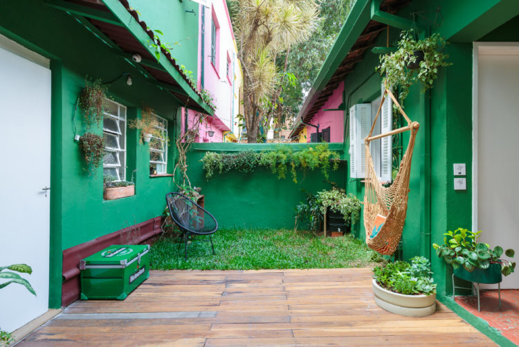 Quintal de casa, espaço aberto entre duas entradas, com todas as paredes externas pintadas de verde.. Um pedaço com grama ao fundo, uma cadeira de balanço suspensa.
