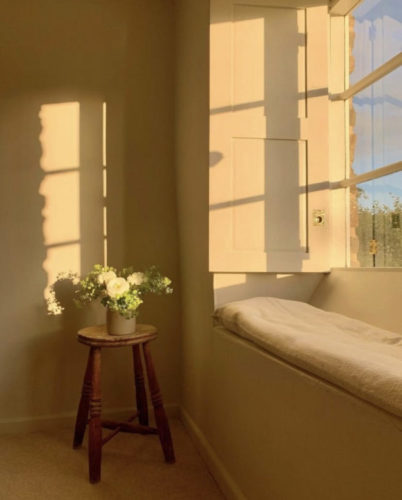 Luz de entardecer pela janela, com um assento embaixo e na frente, um banquinho de madeira com um vaso de flor em cima. 