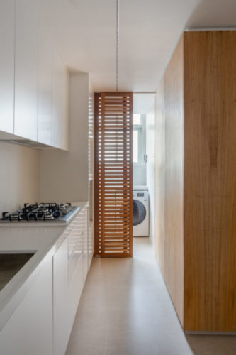 Cozinha toda na cor branca; armários e bancada; treliça vertical em madeira separa a área de serviço.