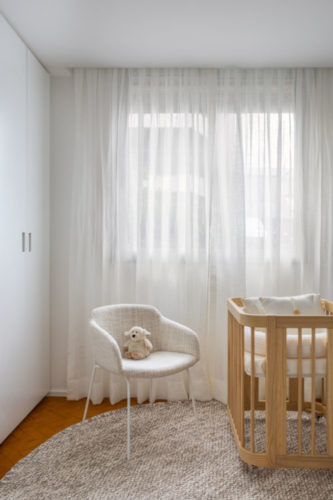 Quarto de bebe com decor. minimalista. Apenas um berço em madeira e ao lado, uma poltrona branca. Ao fundo, cortina em linho branco.