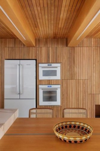Cozinha com paredes e teto revestidos em madeira ripado, e geladeira, forno e micro ondas, todos na cor branca, embutidos.