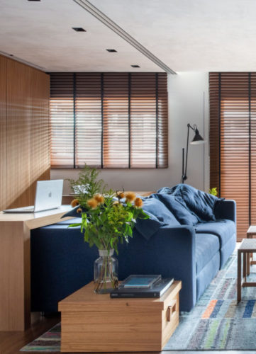 Sala com persianas horizontas em laminas de madeira, sofá azul, tapete em patchwork na cor azul. Atrás do sofá, mesa em madeira serve de home office e a parede de fundo, forrada em madeira. 