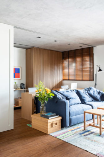 Sala com persianas horizontas em laminas de madeira, sofá azul, tapete em patchwork na cor azul. Atrás do sofá, mesa em madeira serve de home office e a parede de fundo, forrada em madeira. 