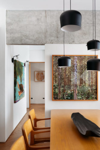 Mesa de jantar e cadeiras em madeira, viga em concreto aparente; nas paredes brancas, telas abstratas coloridas; em cima da mesa pendentes pretos.