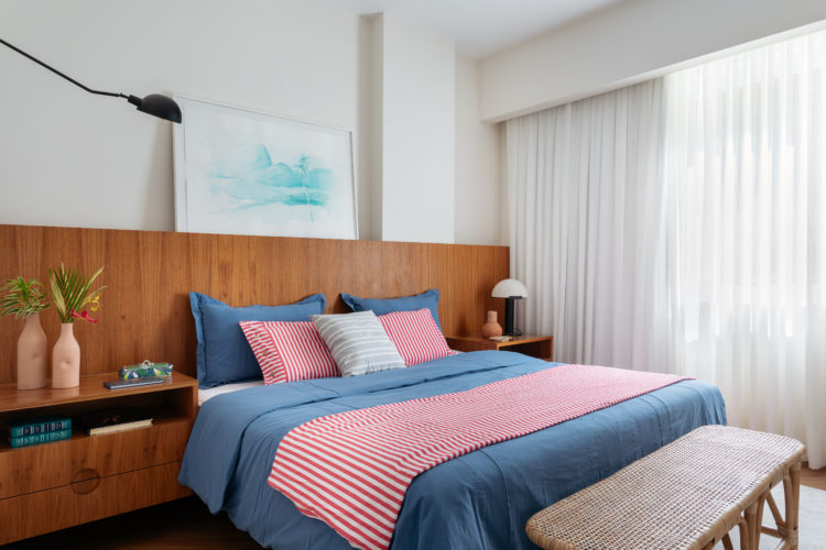 Quarto de casal, com cabeceira em madeira e mesa de cabeceira igual. Enxoval da cama em azul claro e listras de vermelho e branco.