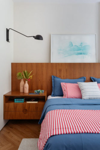 Cabeceira em madeira, aplicada em meia parede, cama com enxoval azul e listra de branco e vermelho.