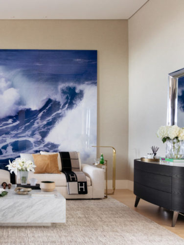 Sala de estar decorada em tons claros, tem como destaque, uma enorme fotografia do mar, apoiada no chão, atrás do sofá. 