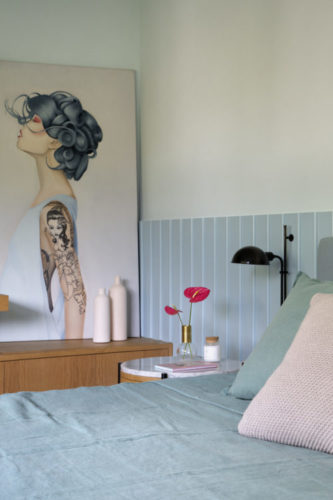 Quarto de casal, com a parede de fundo da cama revestida em meia altura, com ripas de madeira pintadas em azul claro, cabeira da cama em tecido na mesma cor. Ao fundo uma tela com imagem de mulher com o braço tatuado