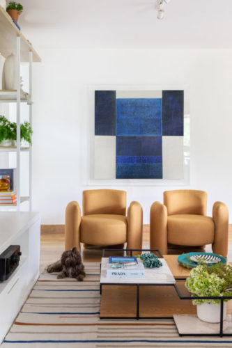 Apartamento decorado em tons claros, duas poltronas em couro claro, e ao fundo, uma tela na cor azul