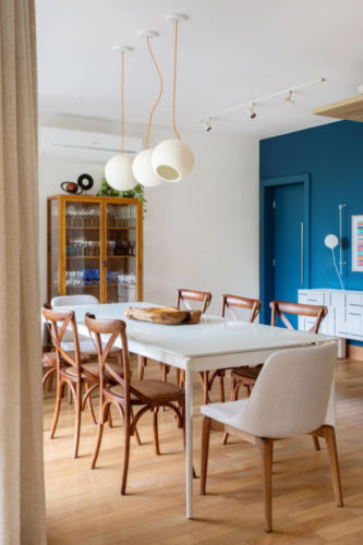 Ambiente de sala de jantar, com mesa com tampo branco, cadeiras em madeira, duas cadeiras brancas nas cabeceiras e ao fundo, parede pintada de azul.