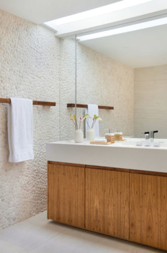 Banheiro com as paredes revestidas com seixos naturais, bancada branca e armário em madeira.
