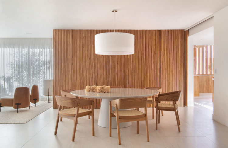 Conforto e acolhimento no apartamento de 450m2 com décor minimalista, ao fundo parede revestida em madeira freijó. Em frente, mesa redonda e cadeiras em madeira