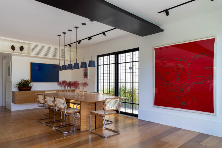 Ambiente sala de jantar. Piso e mesa em madeira, cadeiras em madeira e palinha, uma quadro vermelho na parede