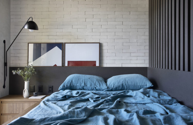 Quarto com parede de tijolinho branco, cabeceira da cama ao longo da parede, em madeira preta, e ao lado da cama, brises em madeira da mesma cor