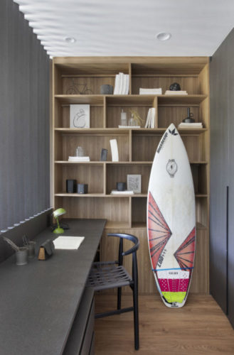 Espaço de estudos no quarto, com bancada em madeira preta, ao lado estante em madeira clara e uma prancha de surf encostada