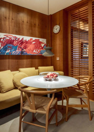 Ambiente de copa na cozinha, com parede de fundo revestida em madeira, sofá amarelo e em frente, uma mesa redonda com tampo de mármore