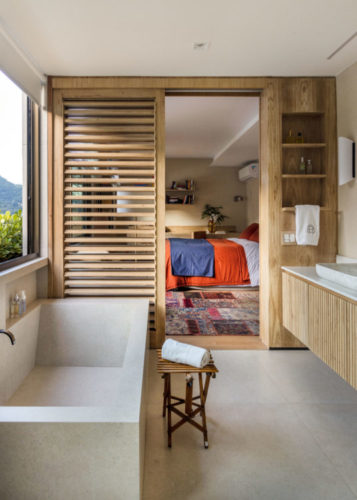 Banheiro com uma banheira e ao fundo o quarto, separado com um brise em madeira