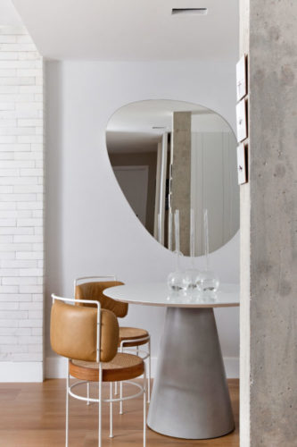 Sala decorada em tons de cinza, com mesa redonda e duas cadeiras de design revestidas em couro. Na parede, um espelho em formato orgânico.