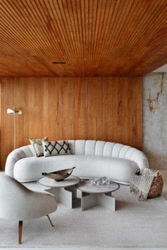 Varanda incorporada a sala, tem paredes e teto revestidos em madeira ripada, um sofá curvo branco, um tapete branco e mesa de centro baixas, decoram o espaço. 