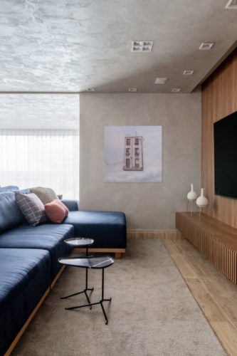 Living com teto e parede pintados com efeito cimento queimado, sofá azul e parede que abriga a tv, forrada em madeira freijó 