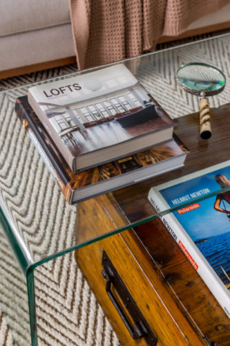 Detalhes de decoração, mesa de cento em vidro, em cima de um baú antigo em madeira, dois livros e uma lupa.
