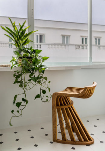 Em frete a janela, que tem vista para a lateral do hotel Copacabana Palace, uma prateleira branca, uma vaso de planta e uma bancada alta em madeira.