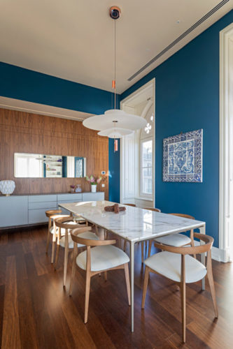 Ambiente de sala de jantar, mesa em mármore, cadeiras em madeira, parede pintada de azul e um painel de azulejos decorando.