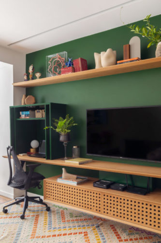 Na sala parede verde com prateleiras madeira e tv na parede. Pequeno armário da mesma cor da parede, com portas camufla o espaço do home office.